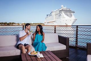 A couple enjoys drinks together on a stylish lounge while the MSC ship is anchored nearby in the Caribbean | MSC Cruises