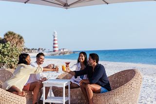 Friends enjoying cocktails together at a beachside table with a lighthouse in the background | MSC Cruises
