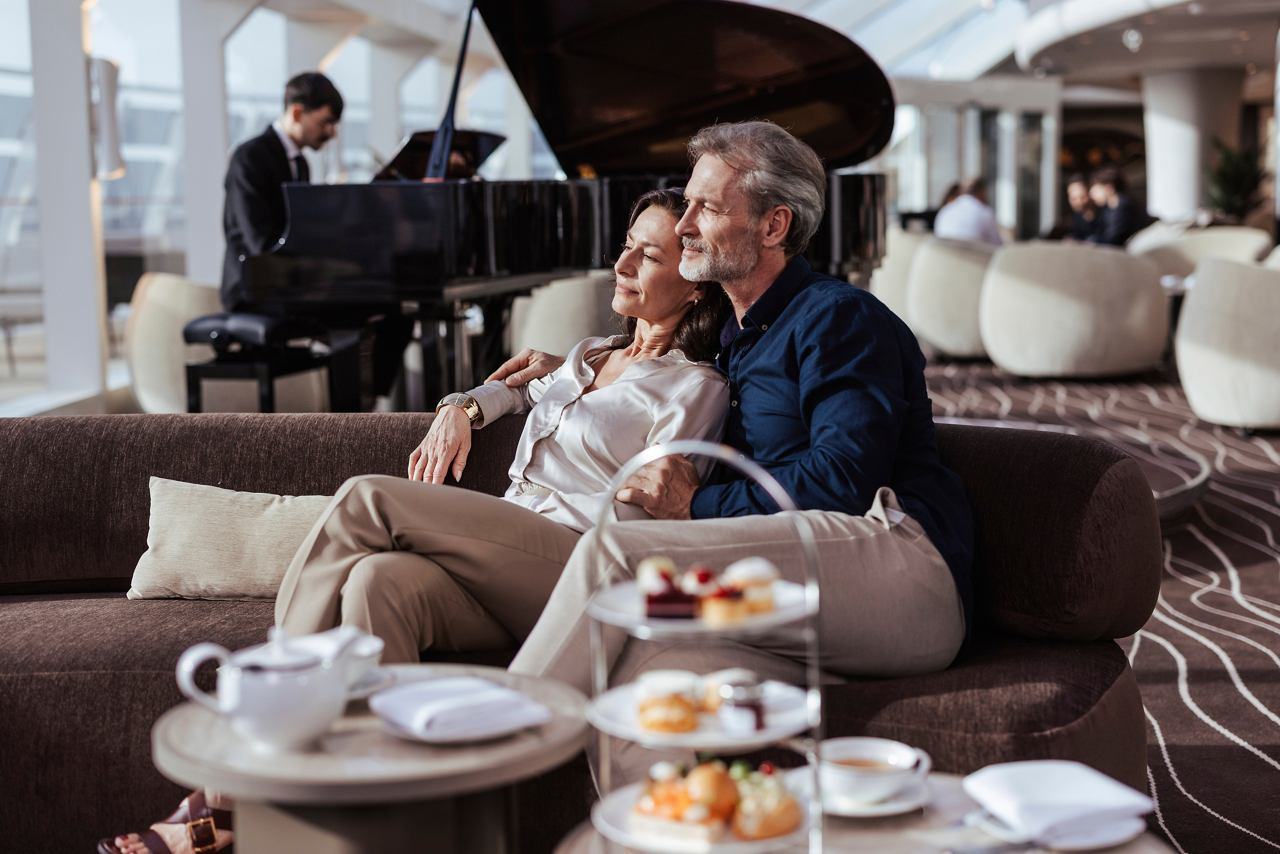 A couple enjoys afternoon tea in the MSC Yacht Club Top Sail Lounge while a musician plays piano nearby | MSC Cruises