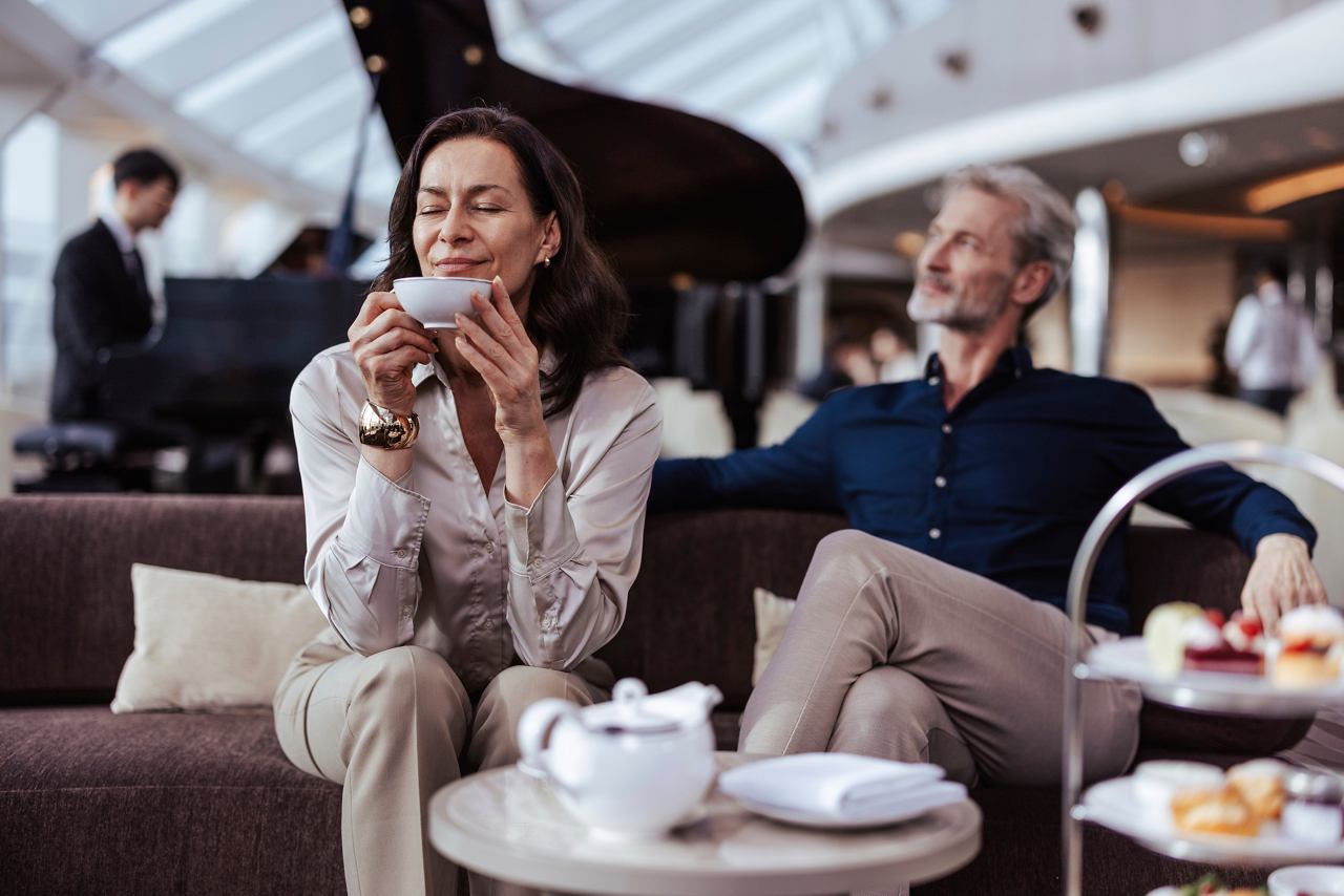 A couple enjoys afternoon tea in the elegant MSC Yacht Club Top Sail Lounge with live piano music in the background | MSC Cruises