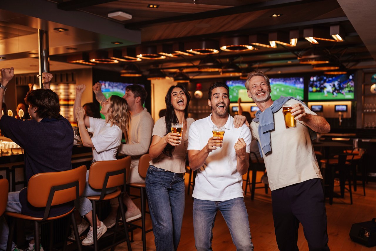 A group of friends cheer together at the bar counter, enjoying drinks in a vibrant sports bar atmosphere | MSC Cruises