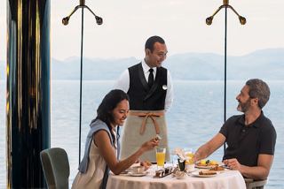 Una pareja disfruta del desayuno sonriendo mientras es atendida por un camarero a bordo. | MSC Cruceros